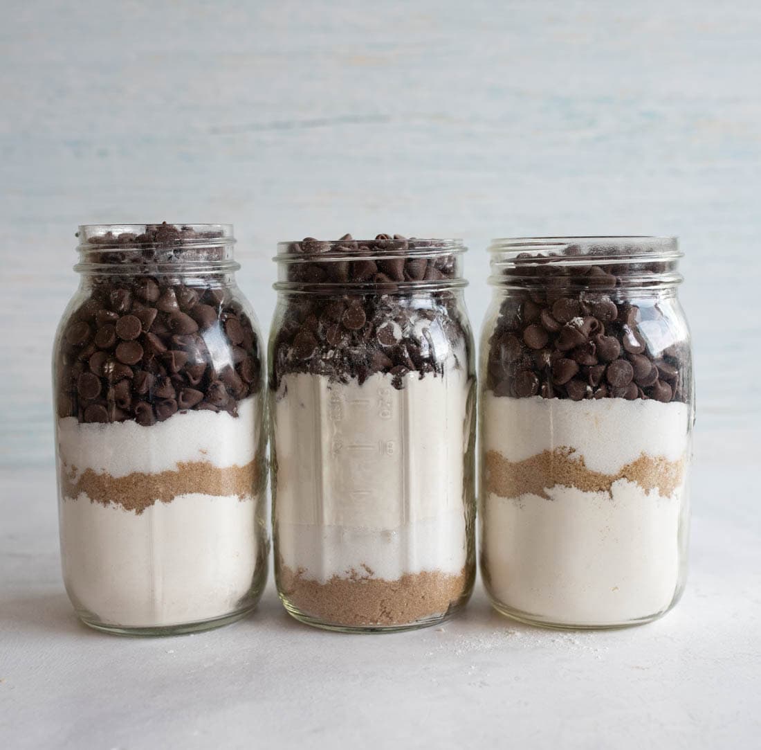 Three glass jars filled with layered ingredients for baking, including chocolate chips, white sugar, and brown sugar. The jars are evenly filled and placed against a light background.