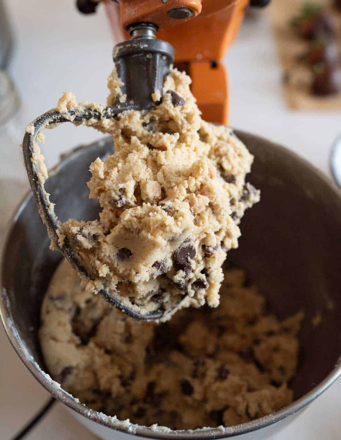 A close-up of cookie dough with chocolate chips on a mixer attachment above a mixing bowl. The dough appears thick and textured. The mixer is orange, and the background shows blurred kitchen items.