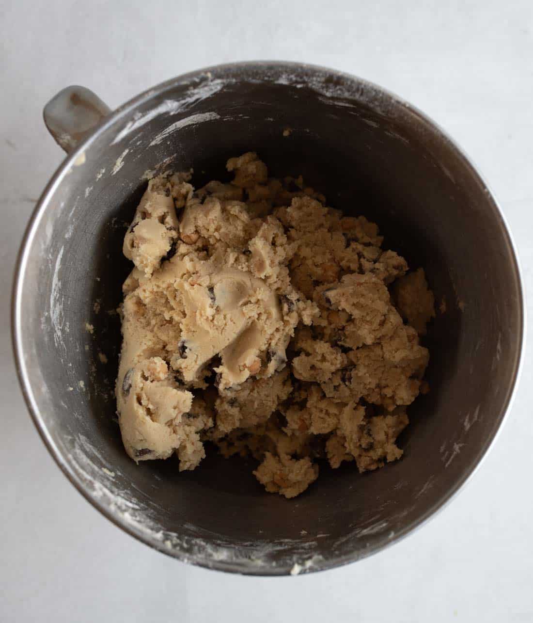 A metal mixing bowl containing unbaked cookie dough with chocolate chips, set on a light gray surface. The dough is slightly uneven, showing a creamy and chunky texture.