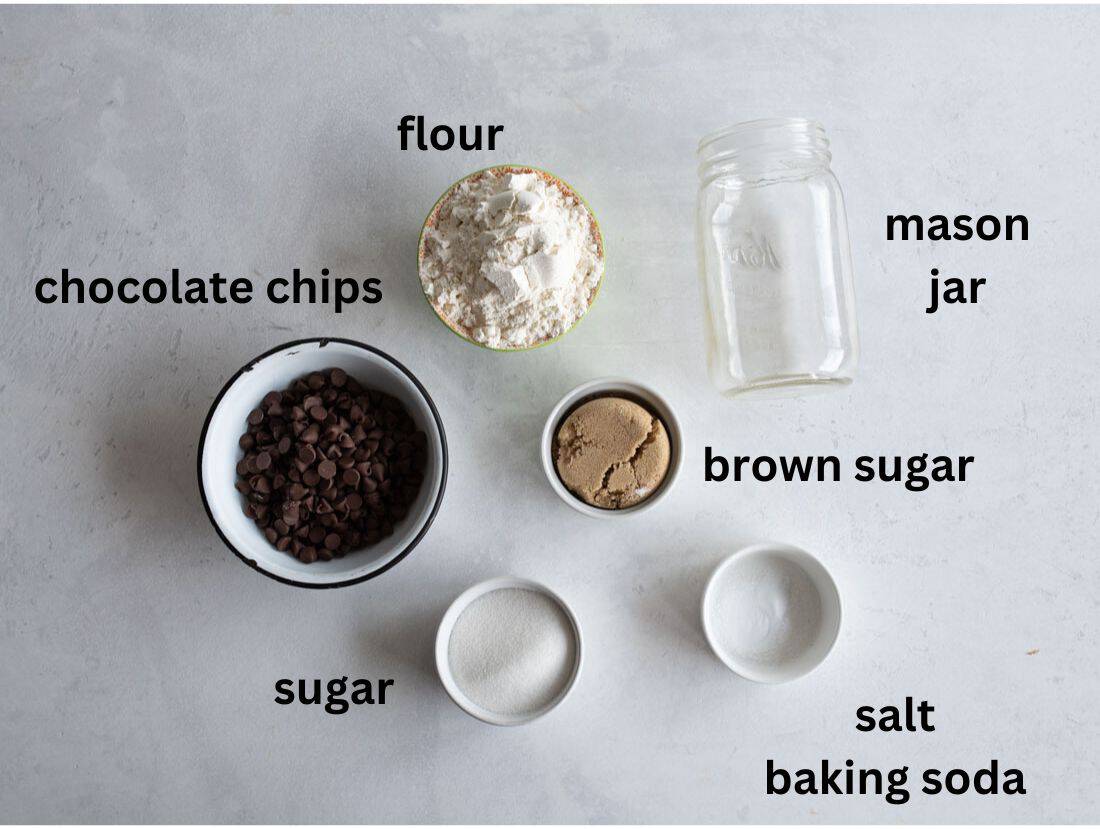 Ingredients for a baking mix laid out on a light surface: a bowl of chocolate chips, a bowl of flour, a jar, a small bowl of sugar, a bowl of brown sugar, and a small bowl containing salt and baking soda.