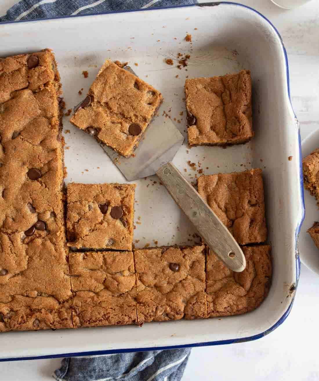 A baking dish with chocolate chip cookie bars, some cut into square pieces. A spatula is lifting a piece from the middle. A few bars are placed on a table nearby. The dish rests on a textured blue cloth.