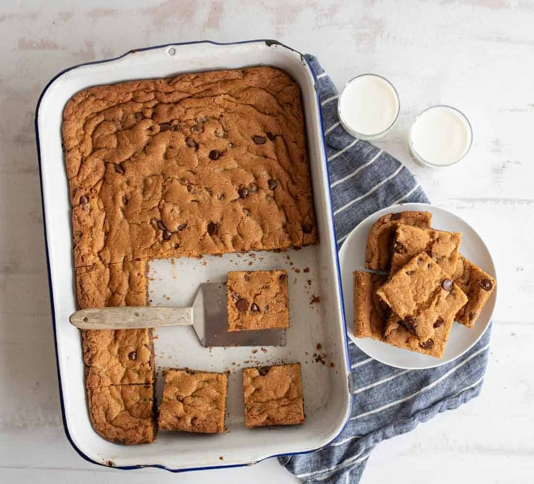 A pan of chocolate chip cookie bars with several pieces cut out and placed on a white plate. The plate rests on a striped cloth, and two glasses of milk are nearby. A spatula is in the pan, next to the cookie bars.