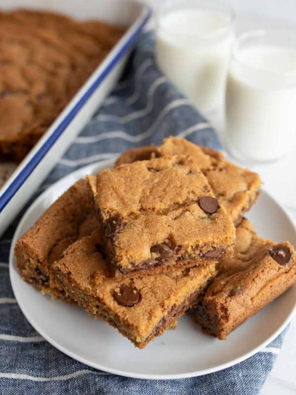 A plate of original Nestle Toll House cookie bars rests on a striped napkin, with more bars in a baking tray nearby. Two glasses of milk are placed in the background, offering a classic pairing.