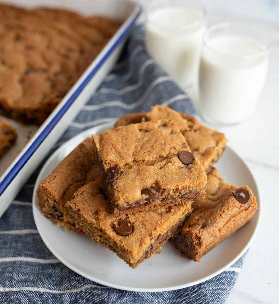 A plate of original Nestle Toll House cookie bars rests on a striped napkin, with more bars in a baking tray nearby. Two glasses of milk are placed in the background, offering a classic pairing.