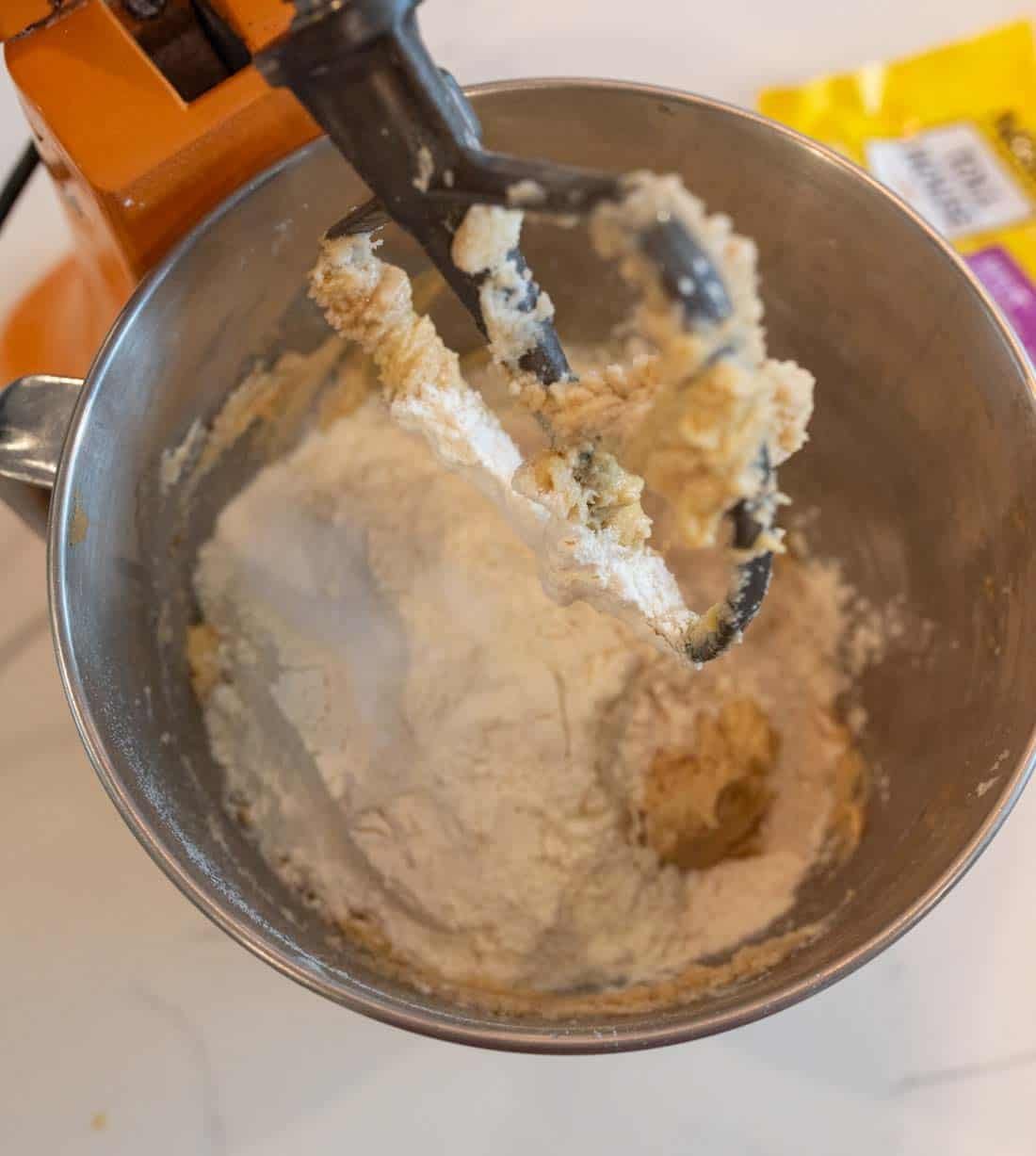 A stand mixer with an orange base is mixing cookie dough. The metal bowl contains a blend of flour and wet ingredients being mixed with a black paddle attachment. A yellow packet is partially visible in the background on a white countertop.