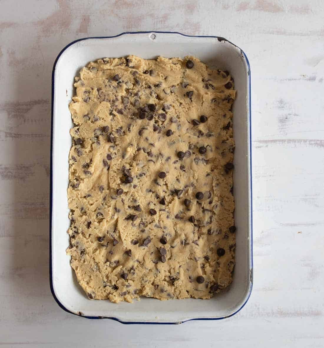 A rectangular baking dish filled with unbaked chocolate chip cookie dough, placed on a light-colored surface. The dough is spread evenly with visible chocolate chips throughout.