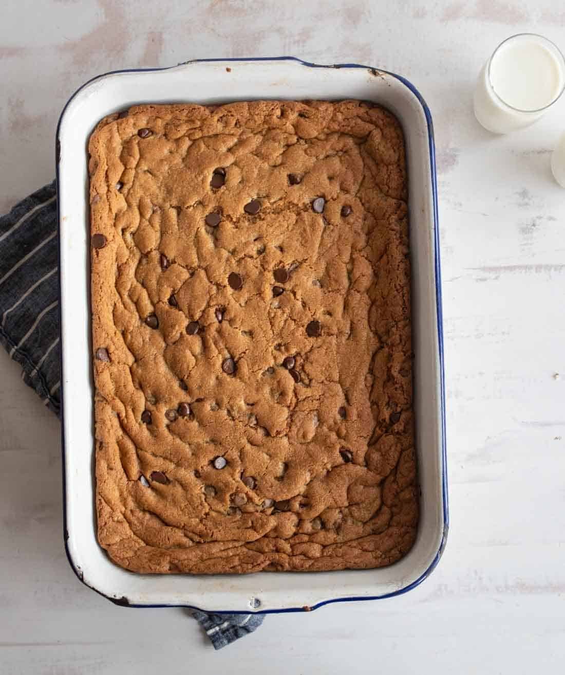 A baking dish filled with freshly baked chocolate chip cookie bars. The edges are golden brown, and the surface is dotted with chocolate chips. A glass of milk sits nearby on a white surface with a striped cloth slightly visible under the dish.