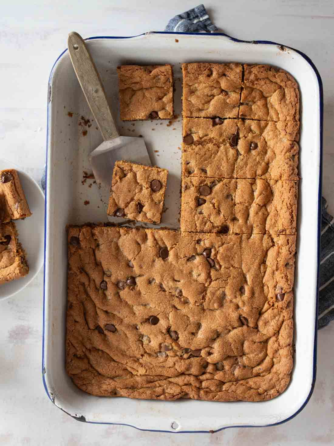 A large pan filled with sliced chocolate chip blondies. One blondie is being lifted with a silver spatula. A small stack of blondies is on a white plate to the side, and a blue cloth peeks out from under the pan.