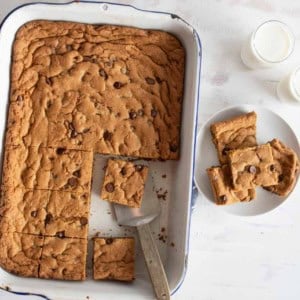 A baking dish with sliced chocolate chip cookie bars. Several bars are served on a white plate beside the dish. Two glasses of milk sit nearby. A spatula rests in the baking dish. The surface is light and textured.