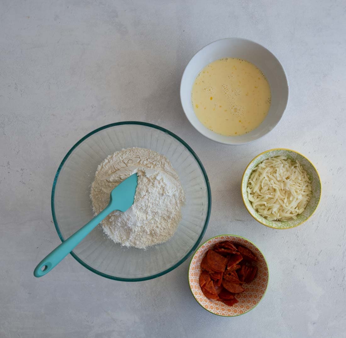 A top-down view of pizza ingredients: a glass bowl with flour and a teal spatula, a bowl with beaten eggs, a bowl of grated cheese, and a bowl of pepperoni slices on a light gray surface.