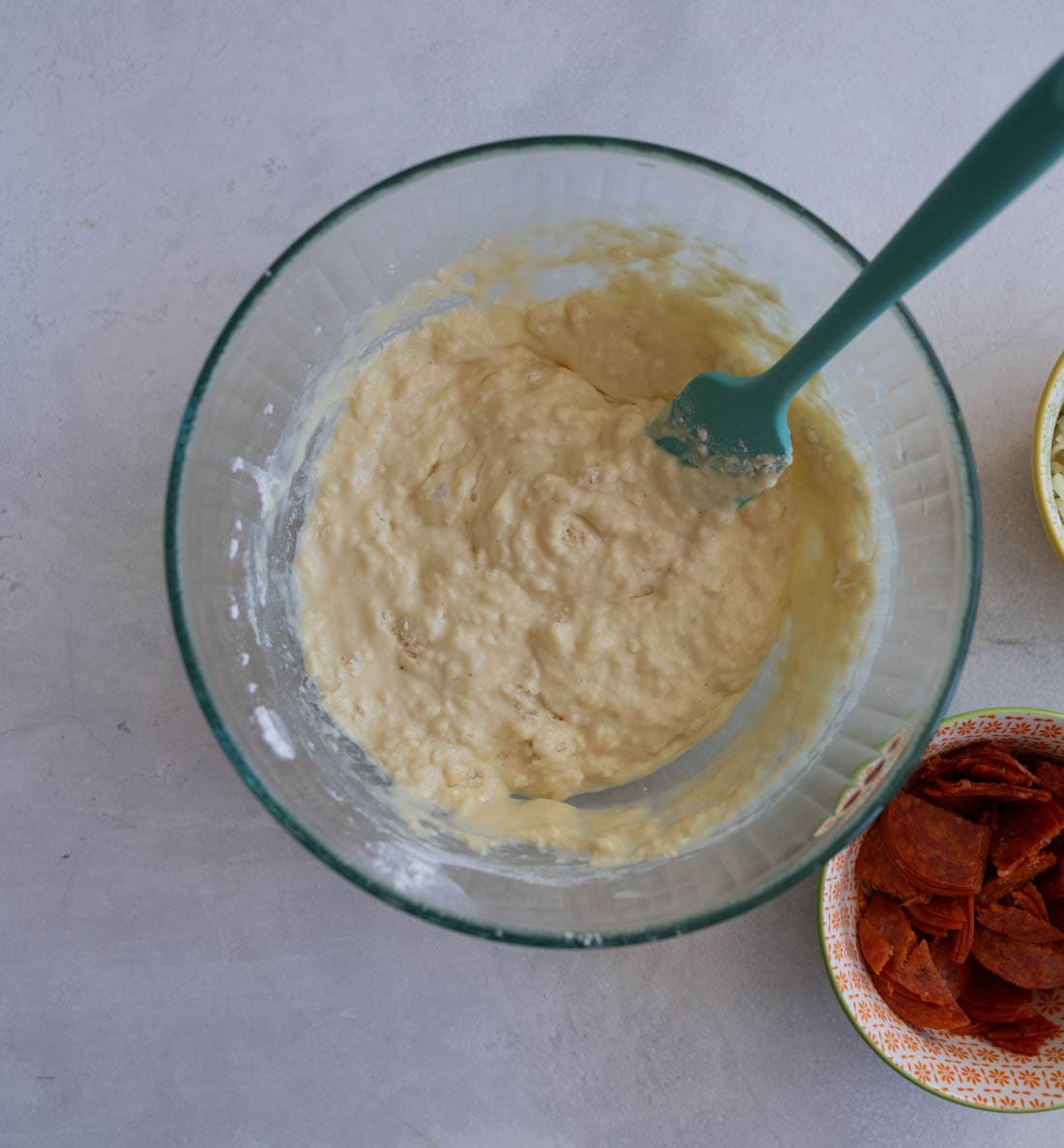 A glass bowl with a mixture of dough and a green spatula inside. Nearby are two small bowls: one with sliced pepperoni and another with grated cheese. The surface is light gray.