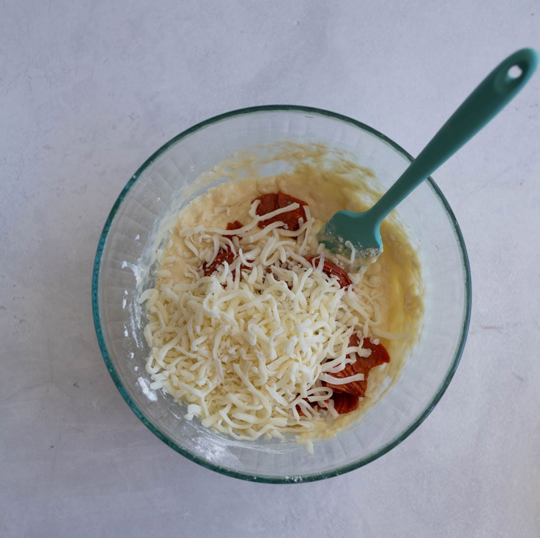 A glass bowl filled with mixed dough, topped with shredded cheese and slices of pepperoni. A teal spatula is partially submerged in the mixture, all set on a light gray surface.