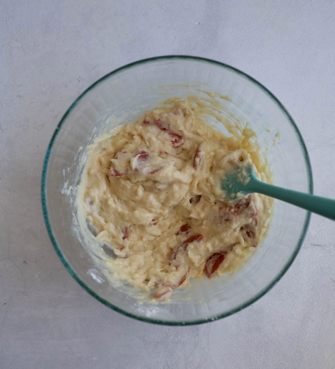 A glass bowl filled with chunky banana bread batter, with visible pieces of banana and other ingredients. A teal spatula is partially submerged in the mixture, which has a creamy texture. The bowl is on a light gray surface.