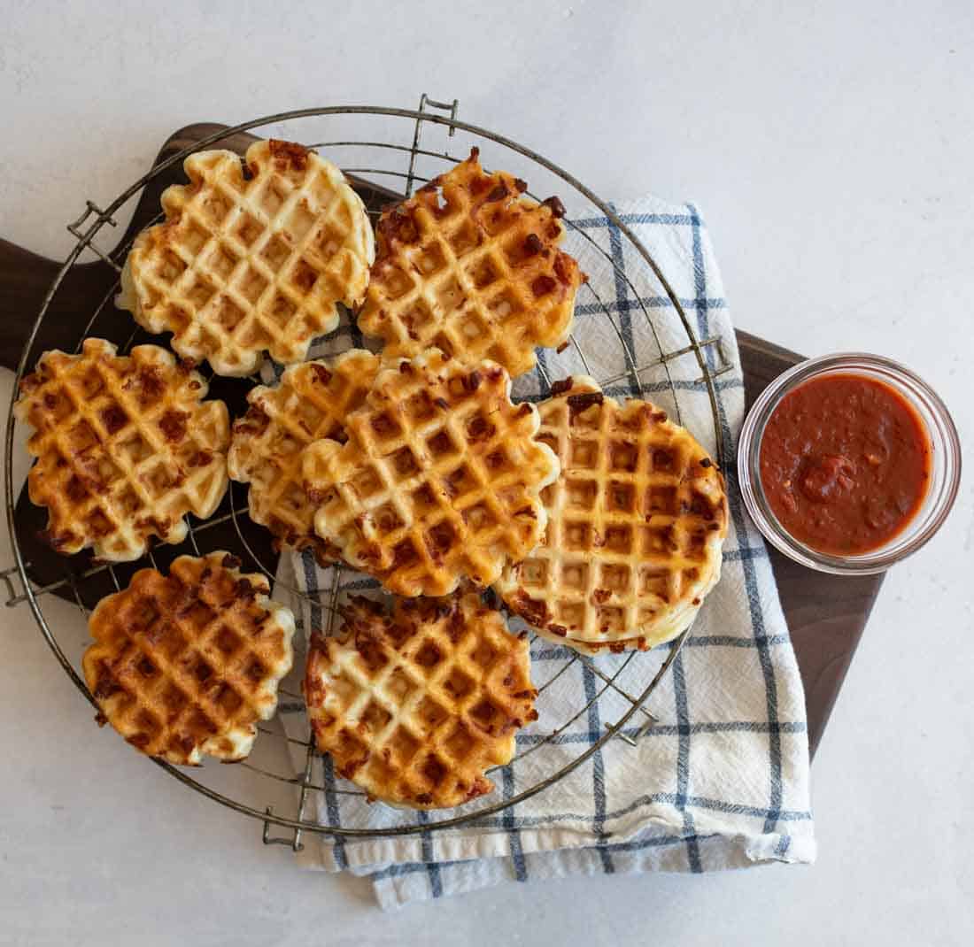 Waffles arranged on a cooling rack atop a wooden board with a checkered cloth. A small jar of red sauce is placed beside them. The waffles are golden brown with a grid pattern.