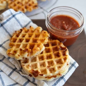 A plate with crispy cheese pizza waffles on a checkered cloth, next to a small jar of red marinara sauce. More waffles can be seen in the background.