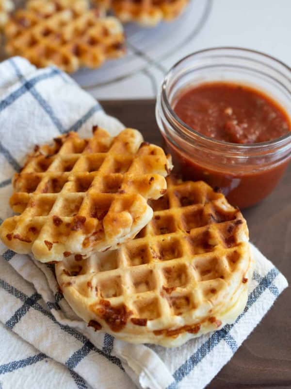 A plate with crispy cheese pizza waffles on a checkered cloth, next to a small jar of red marinara sauce. More waffles can be seen in the background.