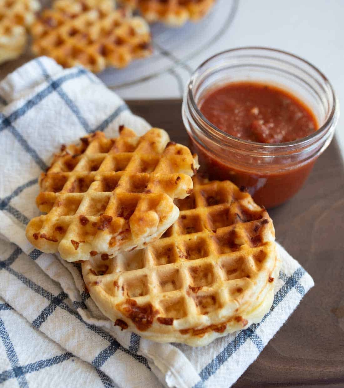 A plate with crispy cheese pizza waffles on a checkered cloth, next to a small jar of red marinara sauce. More waffles can be seen in the background.