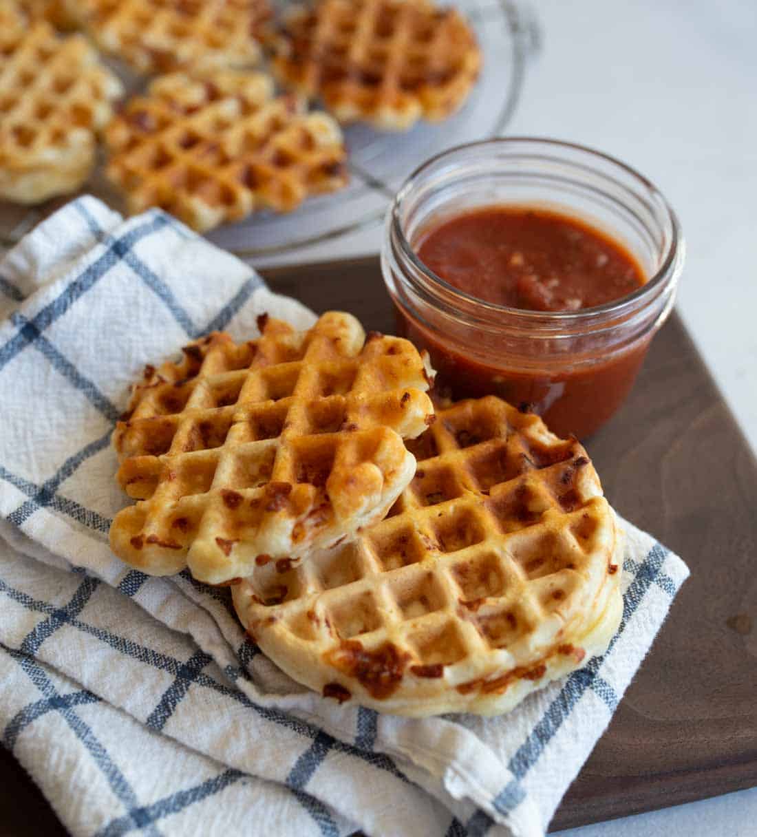 A brown plate holds two waffle pieces next to a small glass jar of red sauce. The waffles rest on a folded blue and white checkered cloth. More waffles are visible on a wire rack in the background.