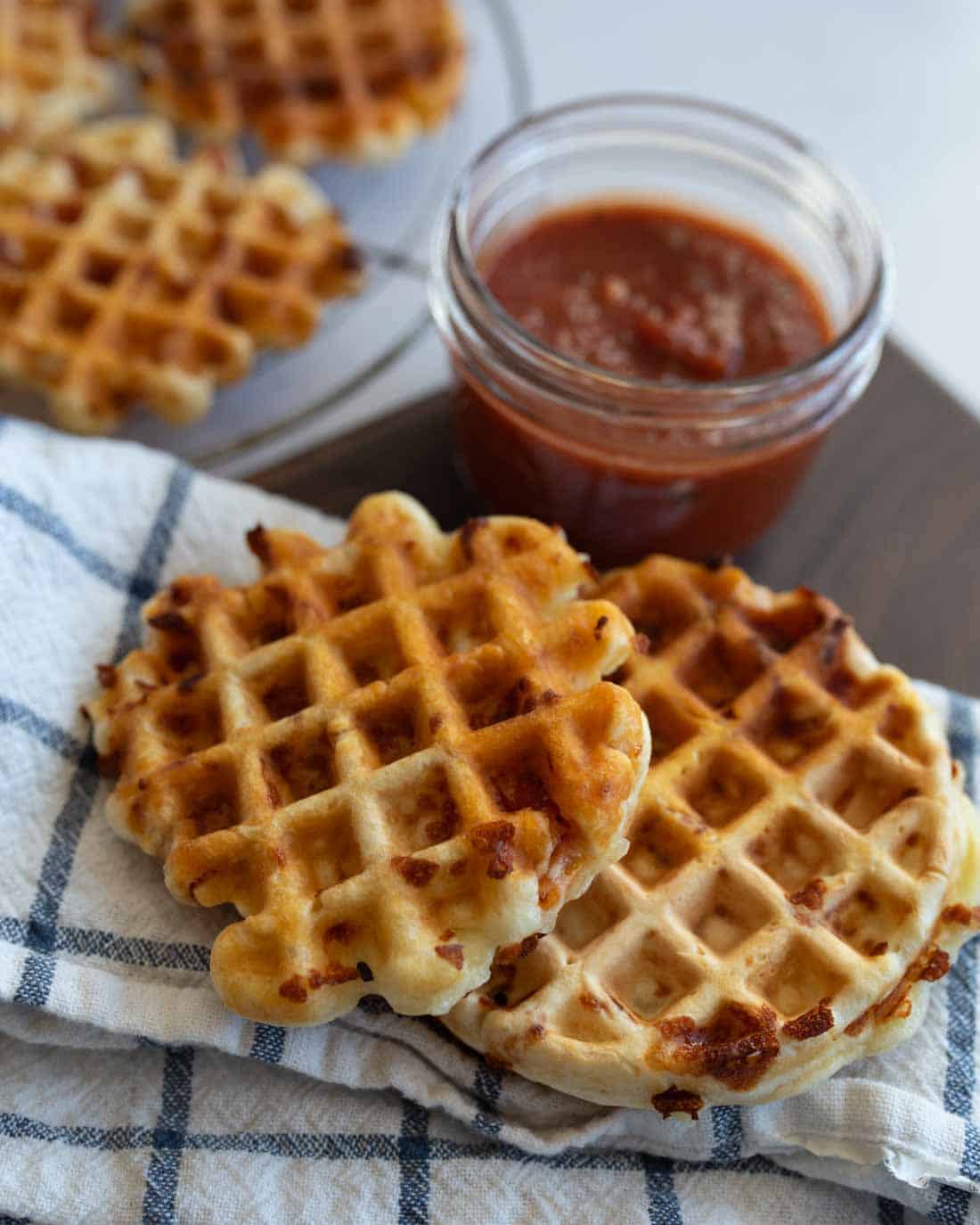 Two golden-brown waffle pieces are placed on a checkered cloth beside a jar of red sauce. More waffles are visible in the background on a plate.