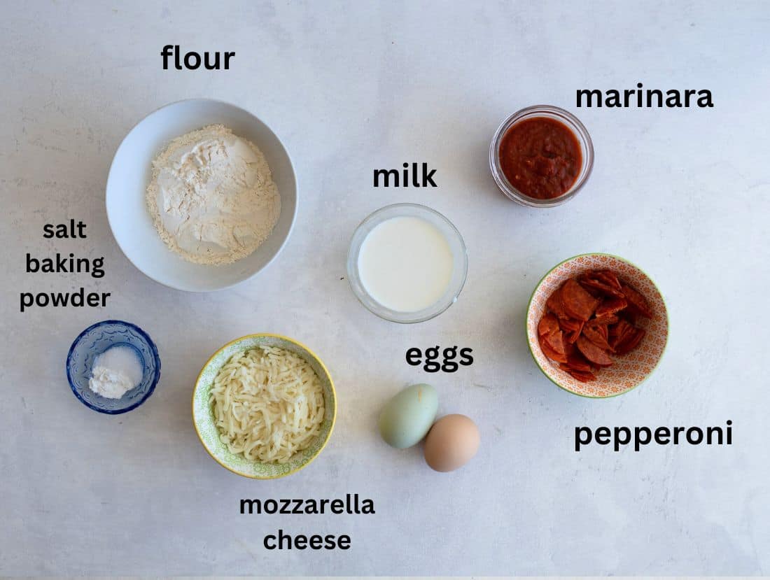 Ingredients for making a dish are arranged on a light surface, including flour, milk, marinara sauce, pepperoni, mozzarella cheese, eggs, and a small bowl with salt and baking powder. Each ingredient is labeled.