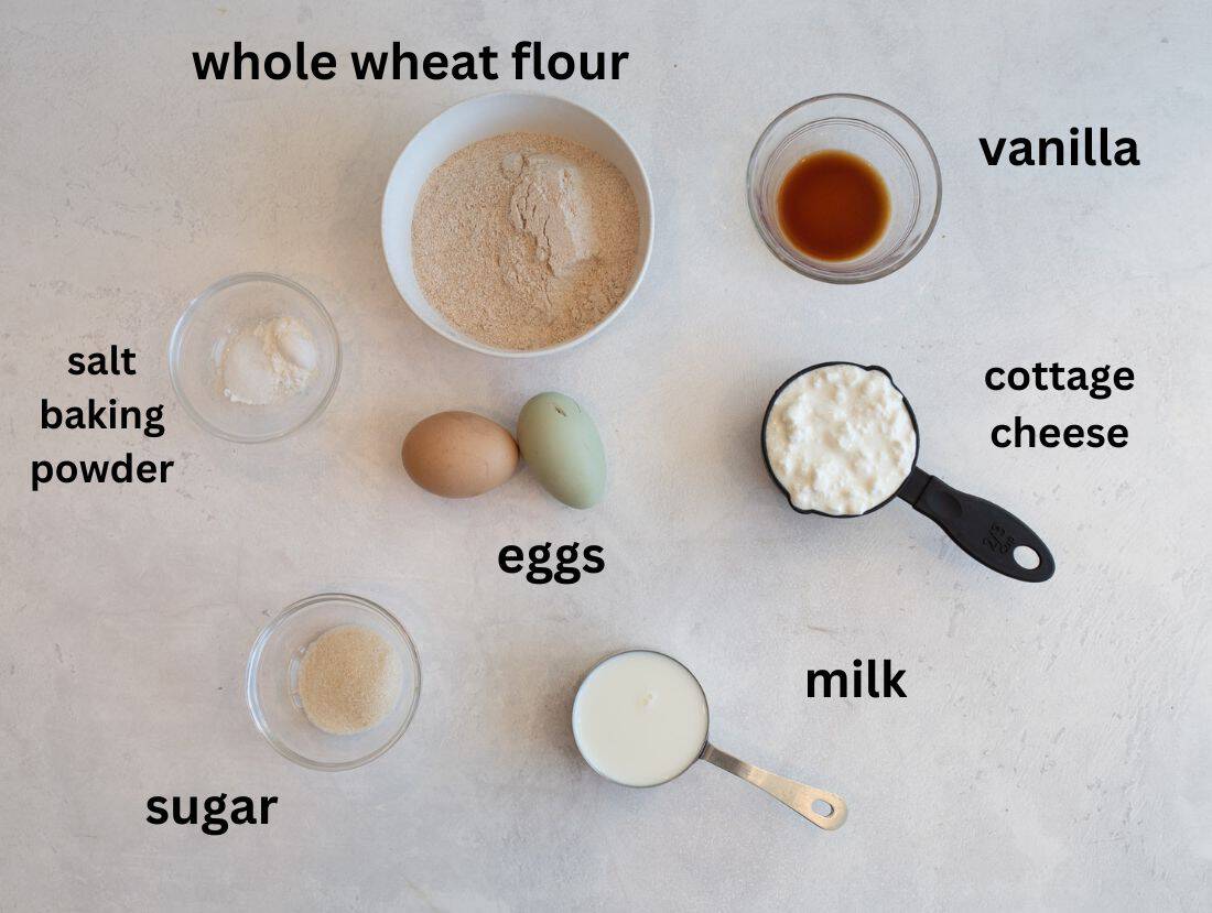 Ingredients for a recipe arranged on a surface include bowls of whole wheat flour, salt and baking powder, sugar, vanilla, cottage cheese, a measuring cup of milk, and two eggs. Each item is labeled with its name.