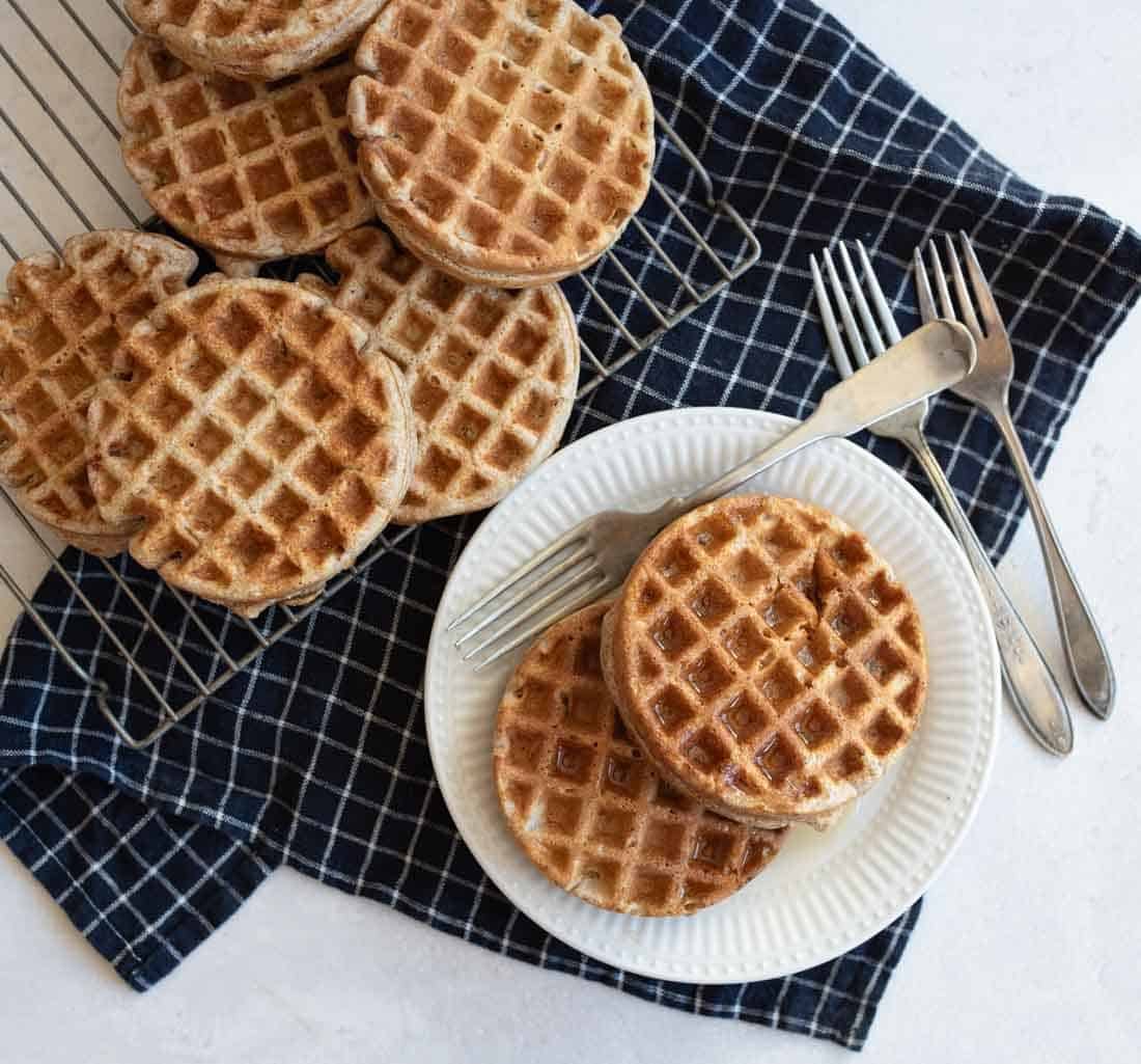 A stack of round waffles on a white plate with a fork, knife, and spoon. More waffles are cooling on a rack beside the plate. The setup is on a dark blue and white checkered cloth on a white surface.