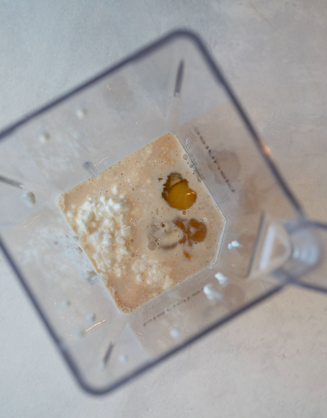 Top view of a blender container with milk, egg yolks, and cottage cheese inside. The ingredients are partially mixed, sitting on a light gray surface.