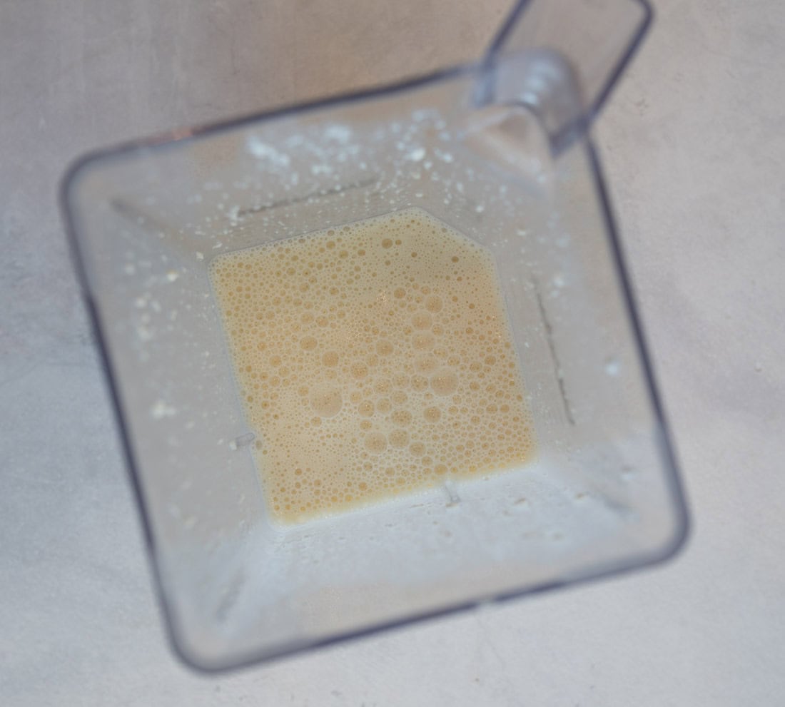 Overhead view of a blender containing a frothy, pale yellow liquid. The blender is placed on a light gray surface, with small bubbles visible on the liquids surface.