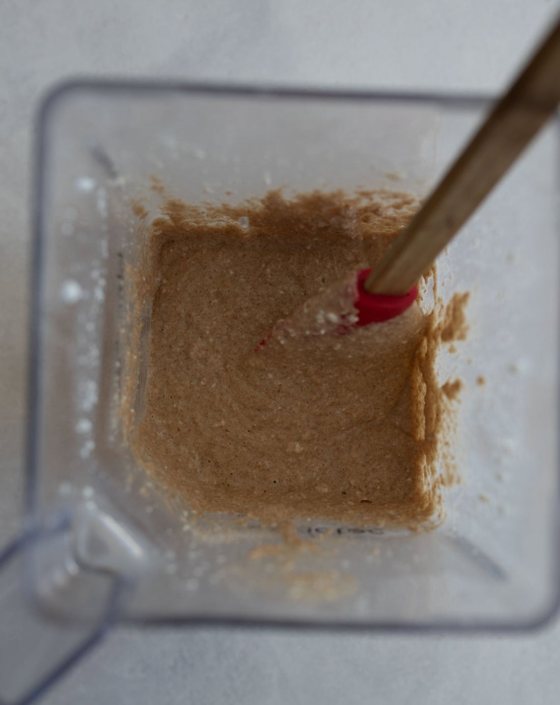 A blender with a thick, light brown batter inside. A red spatula is partially submerged in the mixture, which appears smooth and slightly textured. The blender rests on a light-colored surface.