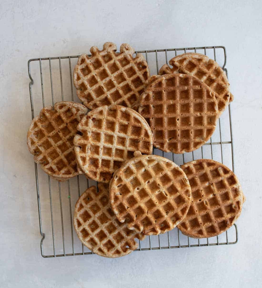 A cooling rack holds nine round, golden-brown waffles with a grid pattern, arranged in a slightly overlapping manner on a light gray surface.