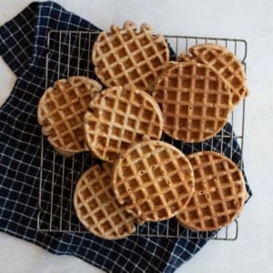 A batch of round waffles is cooling on a wire rack placed over a dark blue checkered cloth. The waffles have a golden-brown color and are arranged in a slightly overlapping pattern on a light countertop.