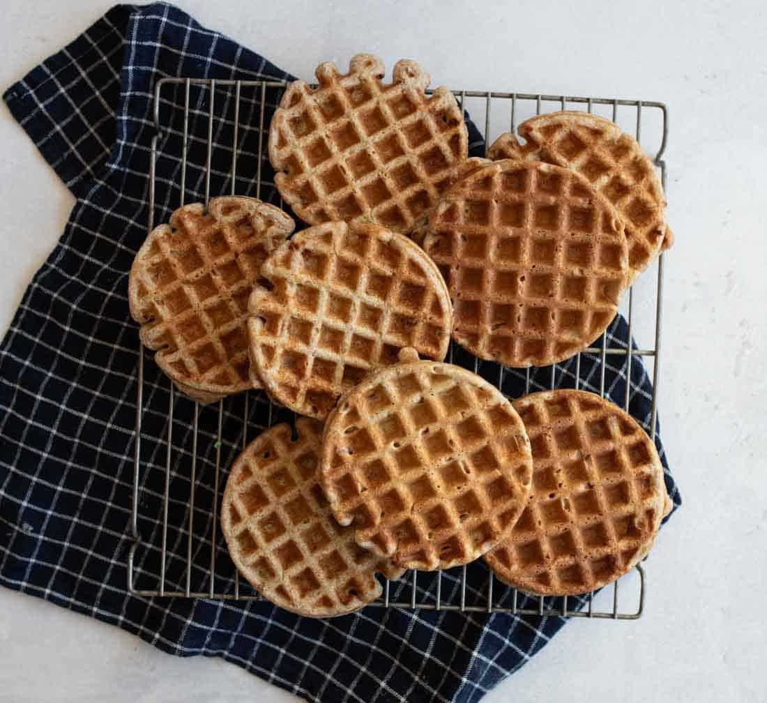 A batch of round waffles is cooling on a wire rack placed over a dark blue checkered cloth. The waffles have a golden-brown color and are arranged in a slightly overlapping pattern on a light countertop.