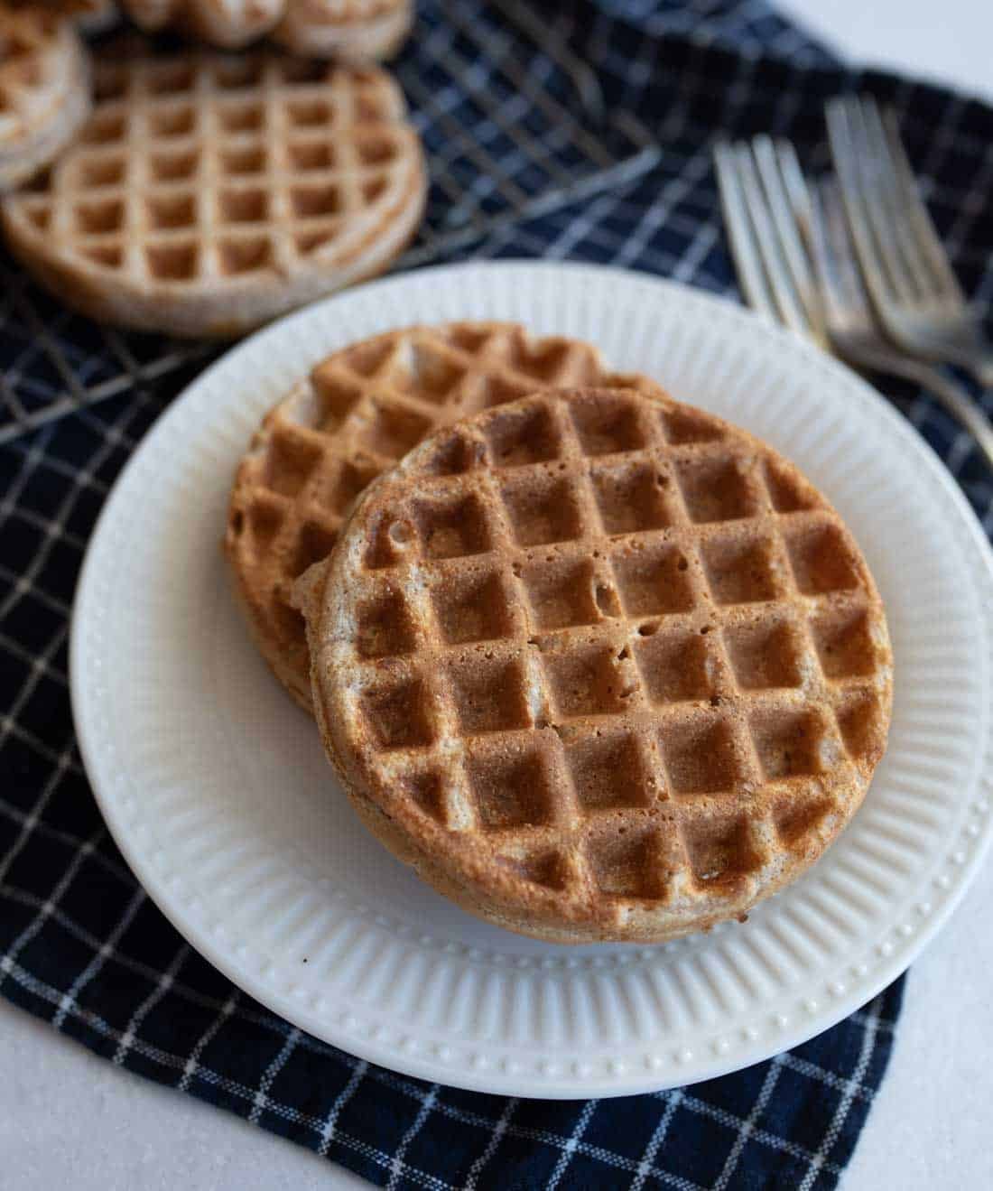 A white plate with two round waffles stacked on it. The plate is placed on a black and white checkered cloth. Two forks sit beside the plate. More waffles are visible in the background.