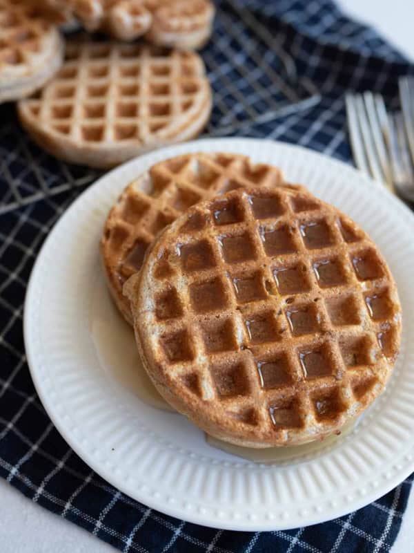 A plate with two round waffles with cottage cheese drizzled with syrup sits on a dark checkered cloth. Two forks are visible to the side. More waffles are partially visible in the background.