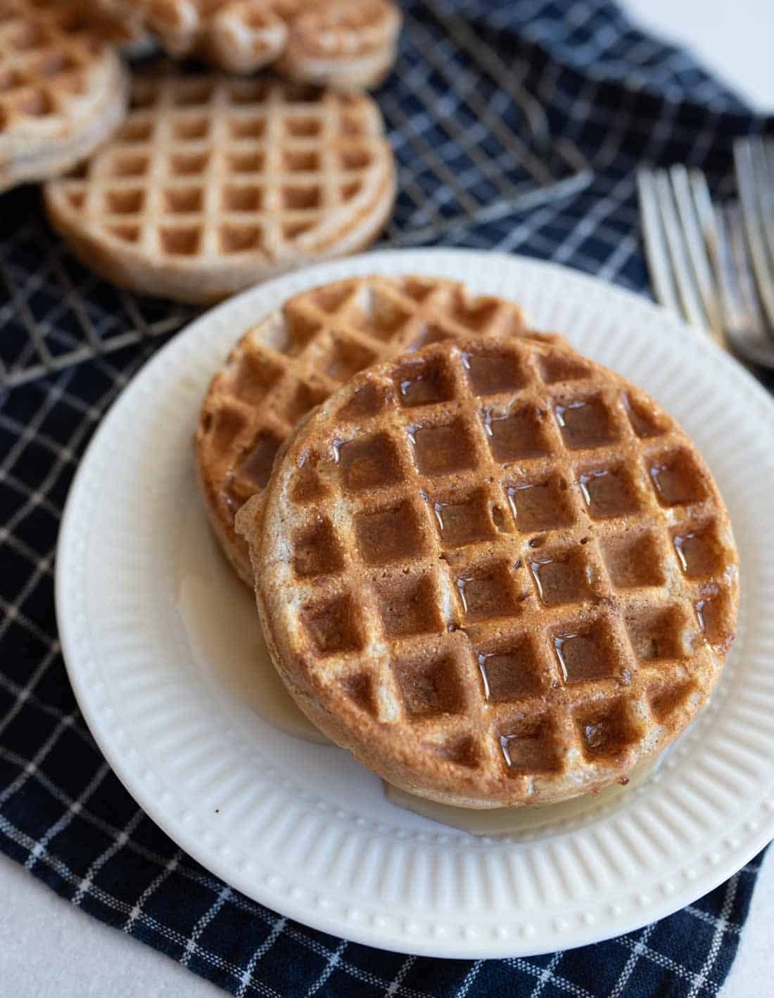 A plate with two round waffles with cottage cheese drizzled with syrup sits on a dark checkered cloth. Two forks are visible to the side. More waffles are partially visible in the background.