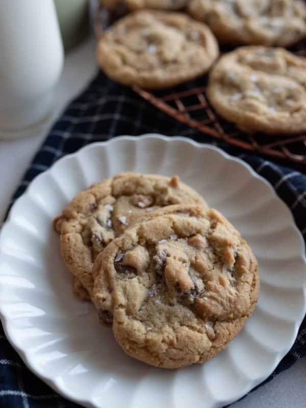 Two chocolate chip cookies are on a white plate, set on a dark checkered cloth. More cookies are visible on a wire rack in the background, with a glass of milk partially visible to the left featuring a salted caramel chocolate chip cookie recipe.