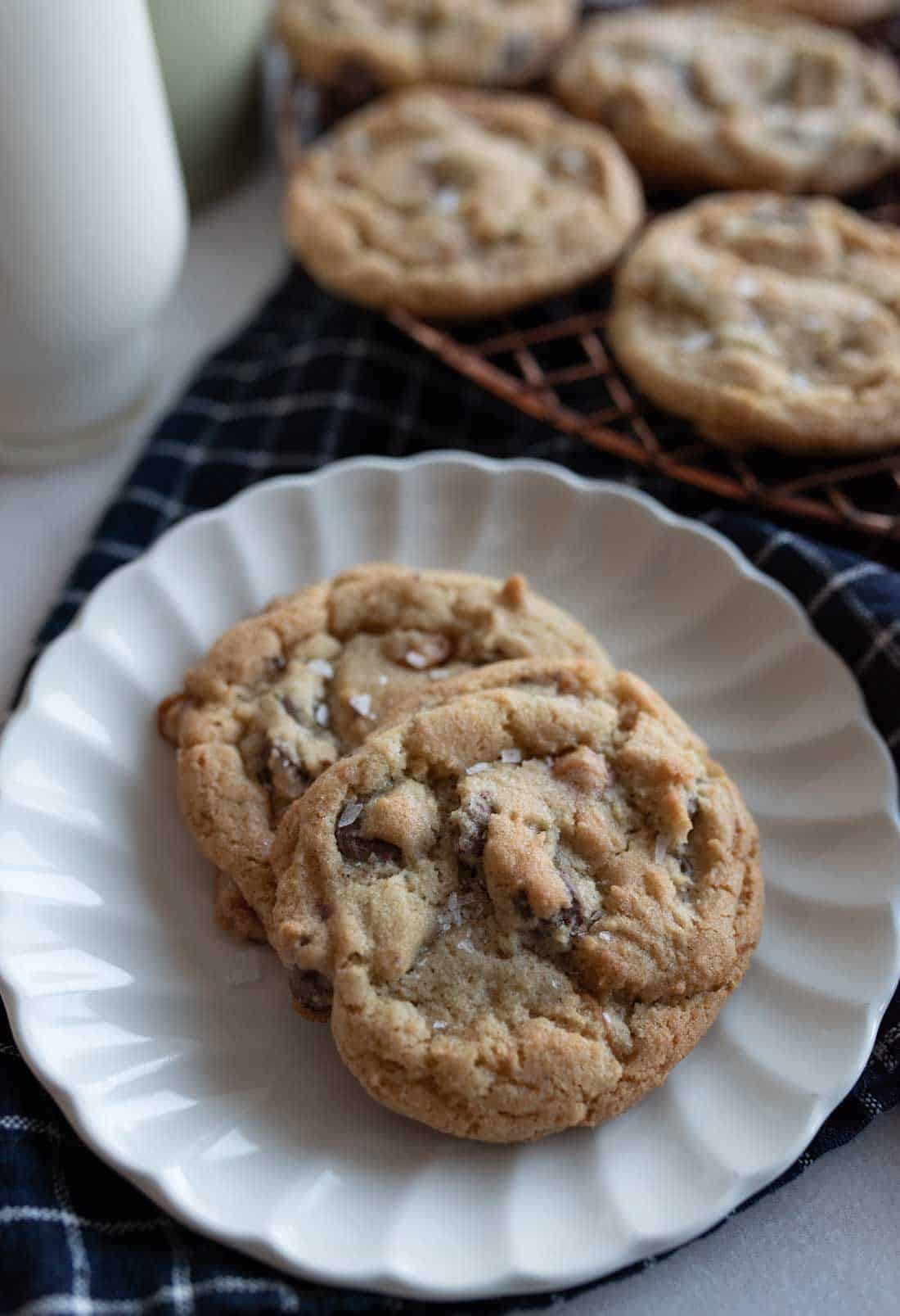 Two chocolate chip cookies are on a white plate, set on a dark checkered cloth. More cookies are visible on a wire rack in the background, with a glass of milk partially visible to the left featuring a salted caramel chocolate chip cookie recipe.