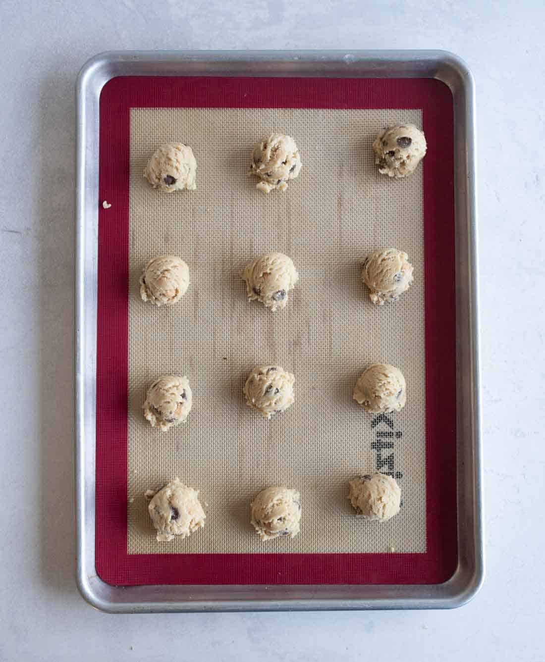 A baking tray holds twelve evenly spaced scoops of cookie dough, placed on a silicone baking mat. The dough appears to contain chocolate chips. The tray is set against a light gray background.