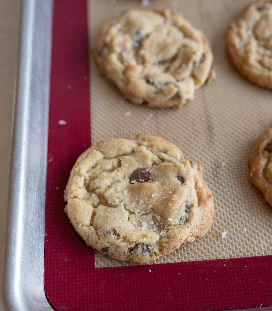 A close-up of chocolate chip cookies on a baking sheet lined with a silicone mat. The cookies have a golden-brown color and are sprinkled with sea salt. One cookie is prominently in focus at the edge of the sheet.