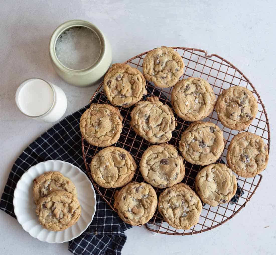 A batch of chocolate chip cookies is cooling on a round wire rack. A glass of milk sits beside them on a black checkered cloth with extra cookies on a white plate. A ceramic jar is partially visible in the background.