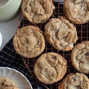 Freshly baked chocolate chip cookies cooling on a round wire rack. A few cookies are placed on a scalloped white plate below. A black and white checkered cloth is underneath, and a coffee cup is partially visible to the side.