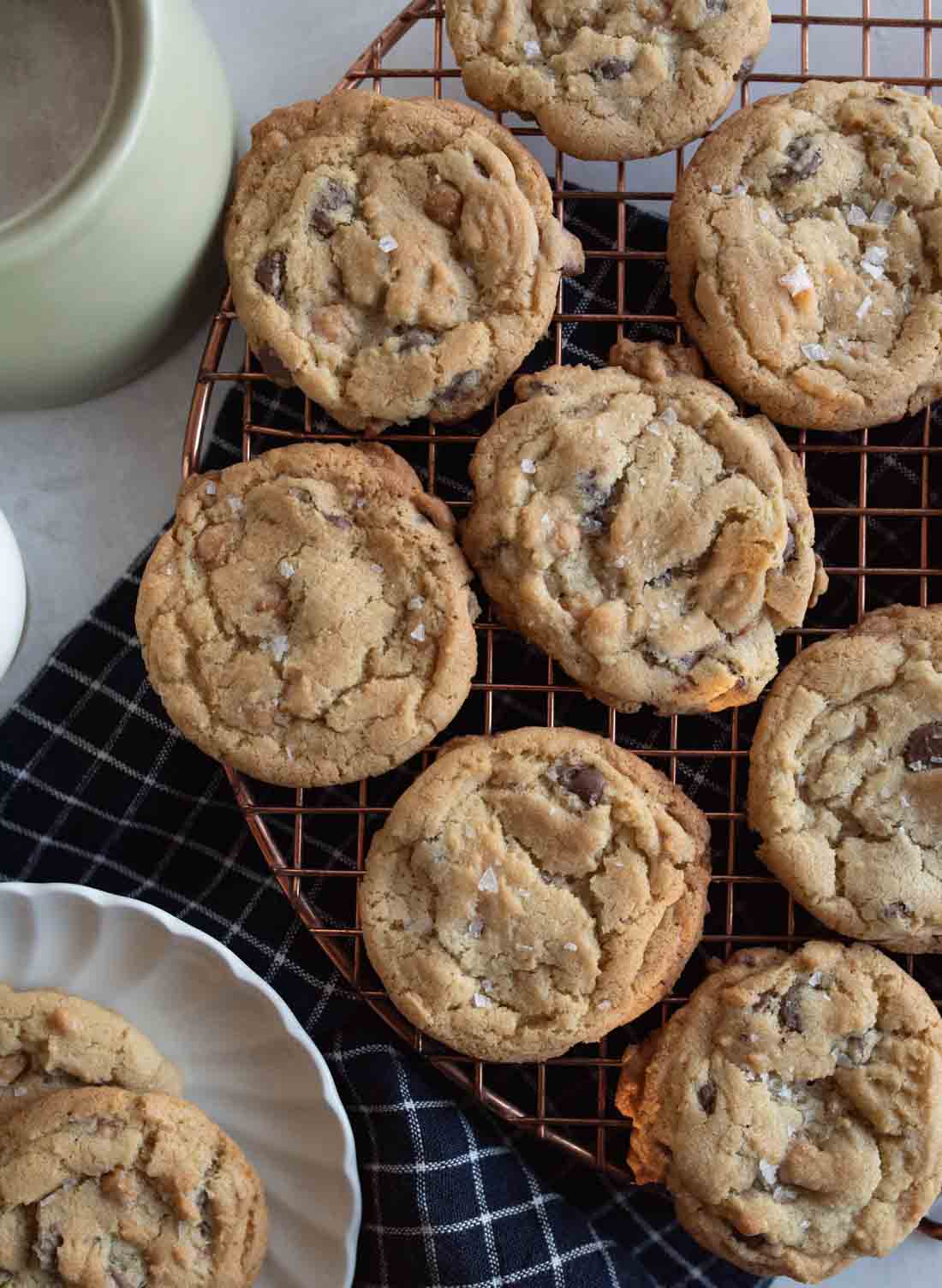 Freshly baked chocolate chip cookies cooling on a round wire rack. A few cookies are placed on a scalloped white plate below. A black and white checkered cloth is underneath, and a coffee cup is partially visible to the side.