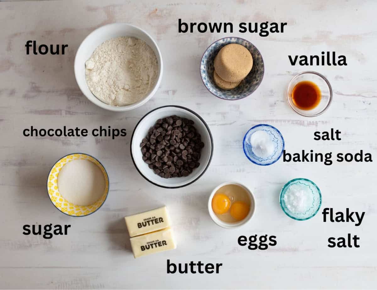 A flat lay of cookie ingredients on a white surface. Labeled bowls contain flour, brown sugar, vanilla, chocolate chips, salt, baking soda, sugar, butter, and eggs. A small dish of flaky salt is also included.