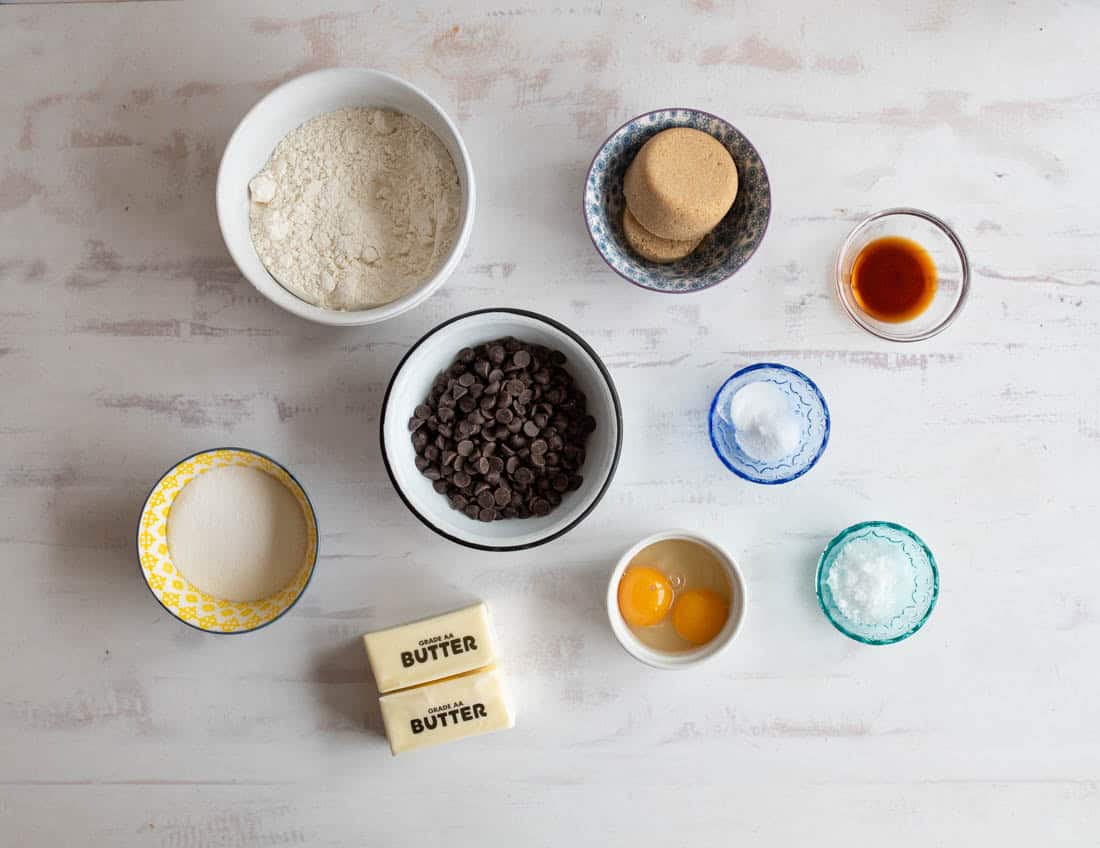 Various baking ingredients arranged on a white surface: flour, chocolate chips, brown sugar, vanilla extract, white sugar, eggs, salt, baking soda, and two sticks of butter.