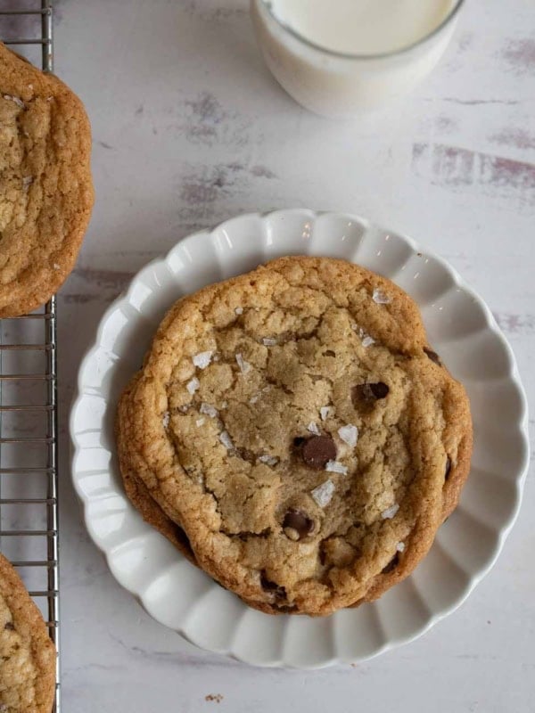 A salty chocolate chip cookie with sea salt flakes graces a white scalloped plate, while part of another cookie rests on a cooling rack. A glass of milk sits in the background on a light, textured surface, completing this delightful scene.