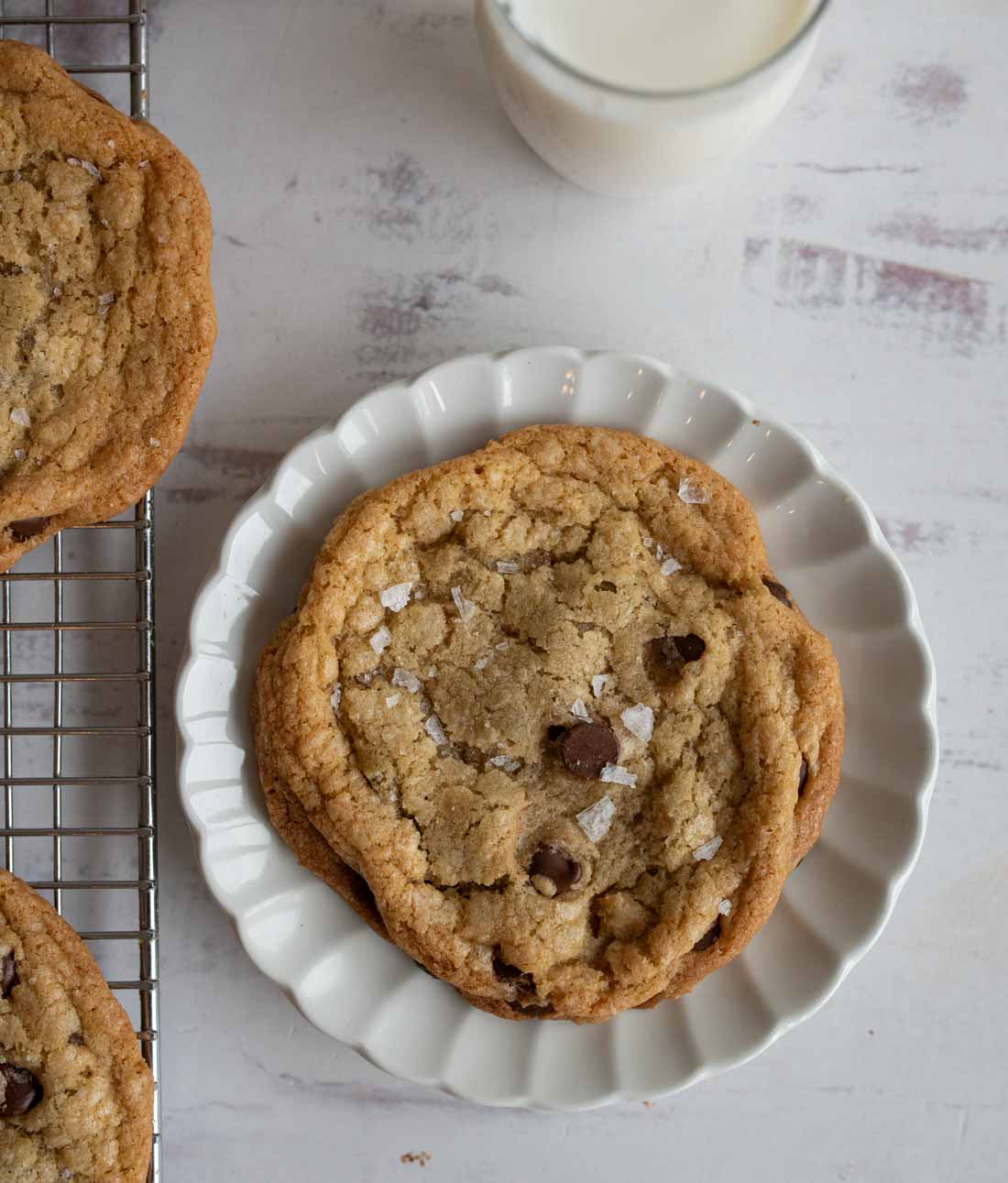 A salty chocolate chip cookie with sea salt flakes graces a white scalloped plate, while part of another cookie rests on a cooling rack. A glass of milk sits in the background on a light, textured surface, completing this delightful scene.