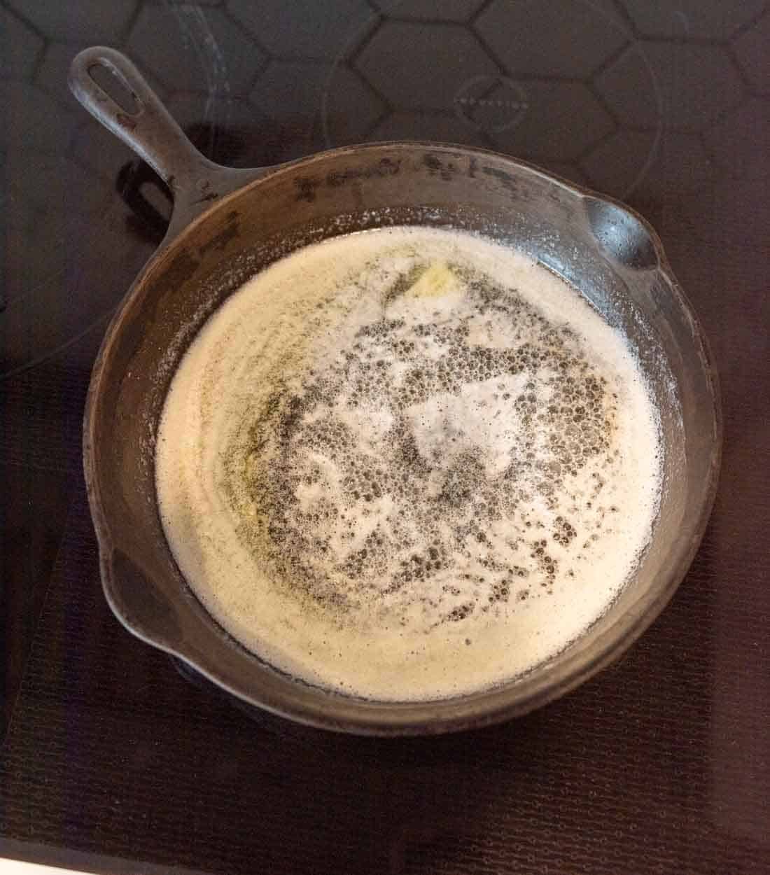 A cast iron skillet on a stovetop contains browning butter. The butter has melted and is bubbling, with small brown specks visible, indicating its reaching the stage of being browned.