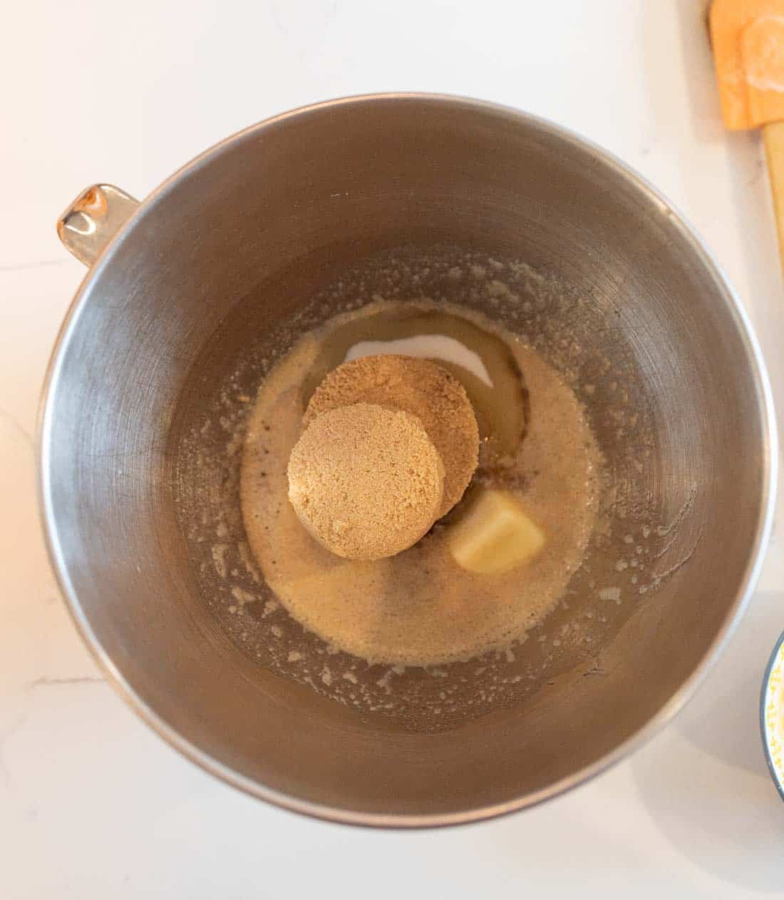 A stainless steel mixing bowl containing melted butter and brown sugar, viewed from above. A spatula with an orange handle is partially visible on the side.