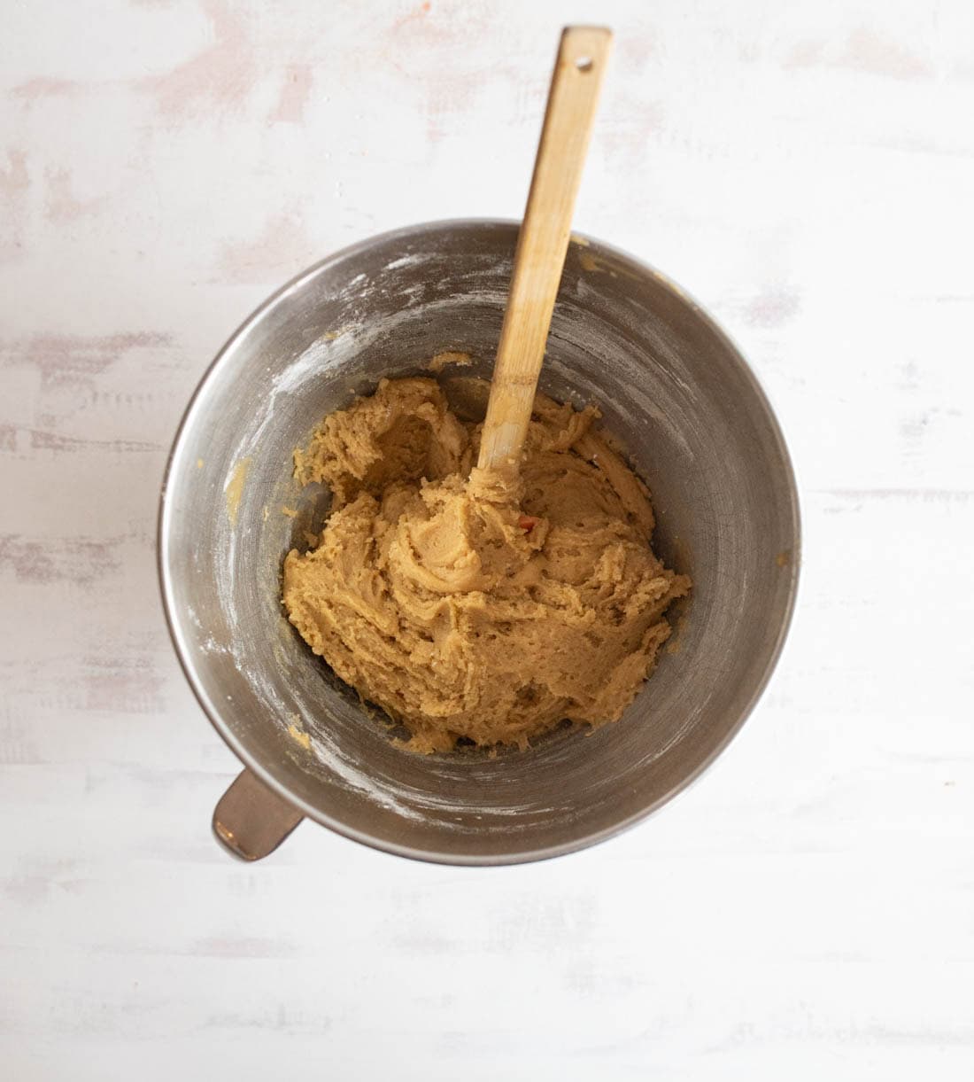 A metal mixing bowl with light brown cookie dough and a wooden spoon on a white textured surface.