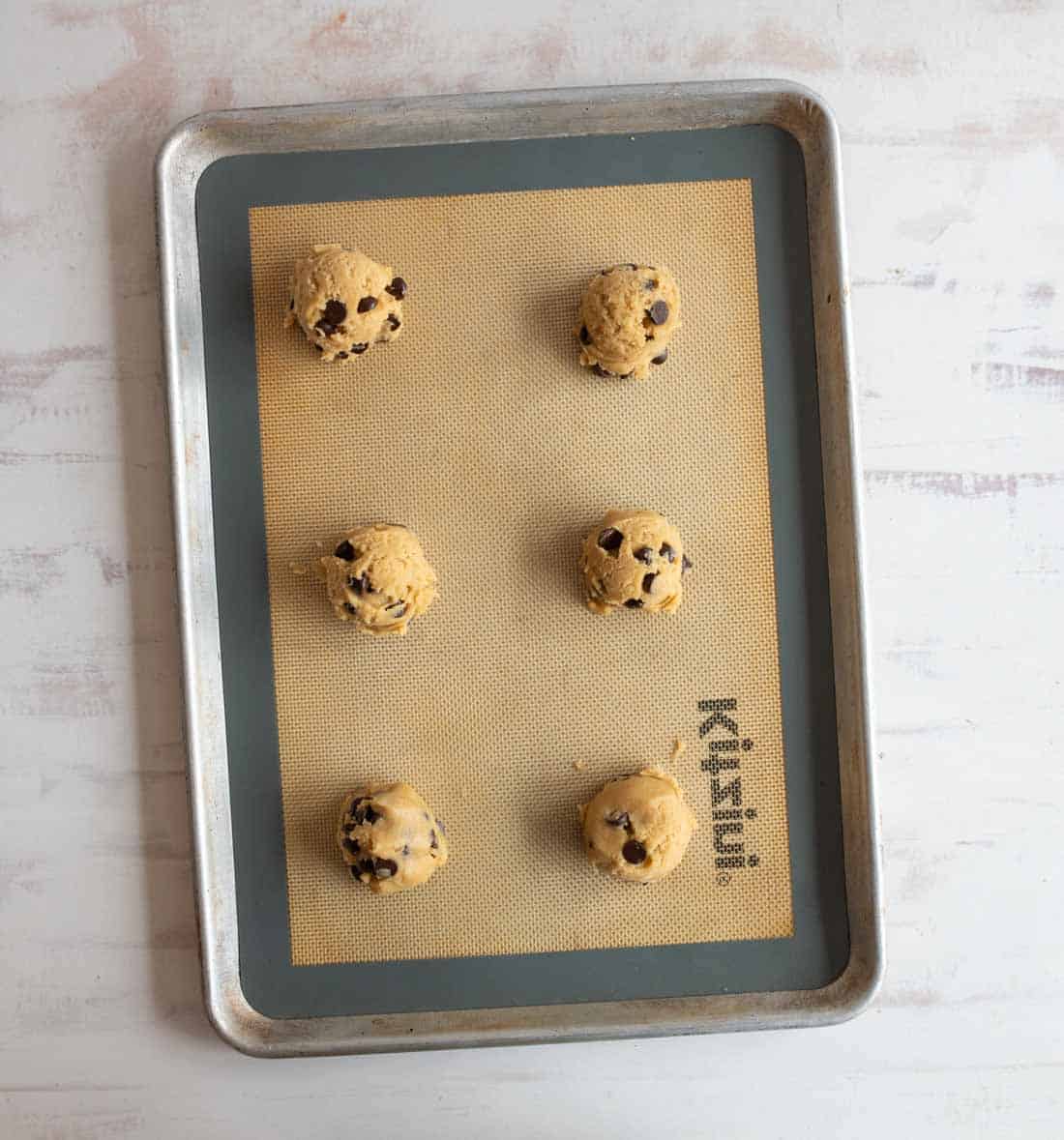 Six balls of chocolate chip cookie dough are arranged on a baking sheet lined with a silicone baking mat. The baking sheet is on a light-colored surface.